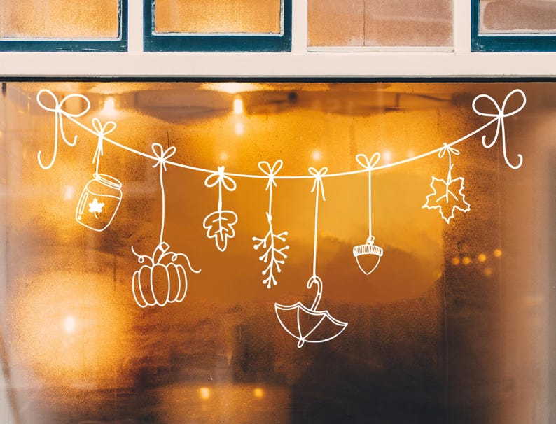 Op de afbeelding: Een witte string met herfstdecoraties hangt voor een raam. De decoraties omvatten een pompoen, een eikel, een pot met een esdoornblad, een tak met bladeren, een tak met bessen en een paraplu.