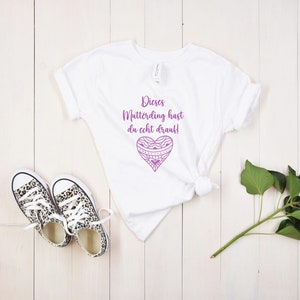 Op de afbeelding: Wit T-shirt met een paars hartontwerp en de tekst "Dieses Mutterding hast du echt drauf!" gedrukt op de voorkant.