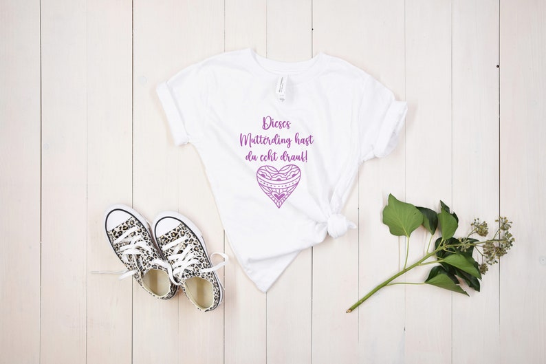 Op de afbeelding: Wit T-shirt met een paars hartontwerp en de tekst "Dieses Mutterding hast du echt drauf!" gedrukt op de voorkant.