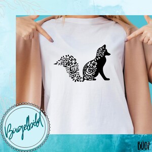 Può includere: Una maglietta bianca con un design di volpe floreale nero. La coda della volpe è fatta di fiori e la volpe è seduta. La maglietta ha il testo "Bügelbild" su un cerchio nell'angolo in basso a sinistra.