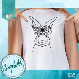 Puede incluir: Una camiseta blanca con un dibujo de línea negra de un conejito con una corona de flores. El conejito está de frente con las orejas levantadas y los ojos cerrados. La corona de flores tiene una variedad de flores y hojas.