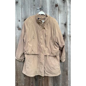 May include: A tan-colored jacket with a high collar and multiple pockets. The jacket has a zipper closure and gold-colored buttons. It is hanging on a white hanger against a wooden background.
