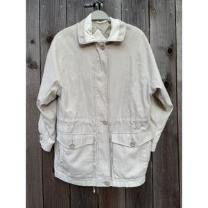 May include: A light beige jacket with a white collar, two front pockets, and button closures. The jacket has a drawstring at the waist and is hanging on a wooden background.