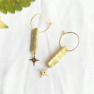 Può includere: Un paio di orecchini a cerchio dorati con pendenti rettangolari in marmo verde chiaro. Ogni pendente è decorato con un piccolo ciondolo a forma di stella dorata a quattro punte che pende dal fondo, creando un design celeste con finitura lucida.