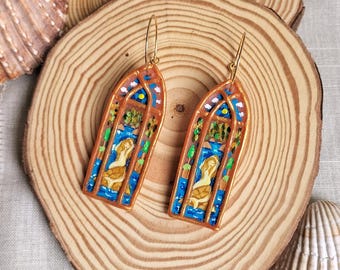 Può includere: Un paio di orecchini pendenti color oro. Ogni orecchino presenta un design a vetrata con una figura di sirena, su sfondo blu e arancione. Gli orecchini sono esposti su una fetta di legno, con conchiglie sullo sfondo.