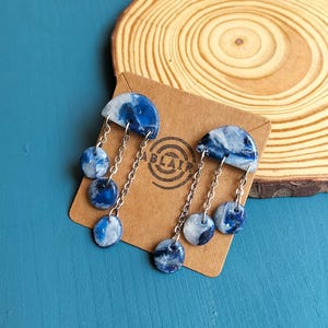 Peut inclure: Boucles d'oreilles en pâte polymère marbrée bleue et blanche. Chaque boucle d'oreille est composée d'un demi-cercle sur le dessus et de trois perles rondes pendantes reliées par des chaînes argentées. Les boucles sont présentées sur une carte marron.