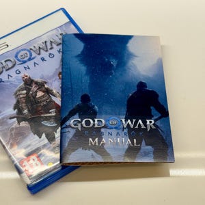 Può includere: Una custodia per giochi PS5 e un manuale per God of War Ragnarök. La custodia del gioco è blu con la copertina di un uomo muscoloso con un'ascia. Il manuale presenta una scena innevata con due figure e il titolo del gioco.