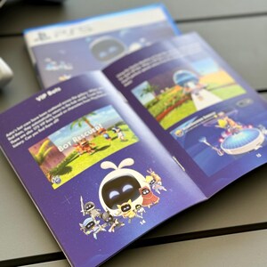May include: An open booklet with a purple cover and a blue background. The booklet features a cartoon character with a white body and a black face. The character is surrounded by other cartoon characters. The text "Bot Rescued!" is visible in the image.