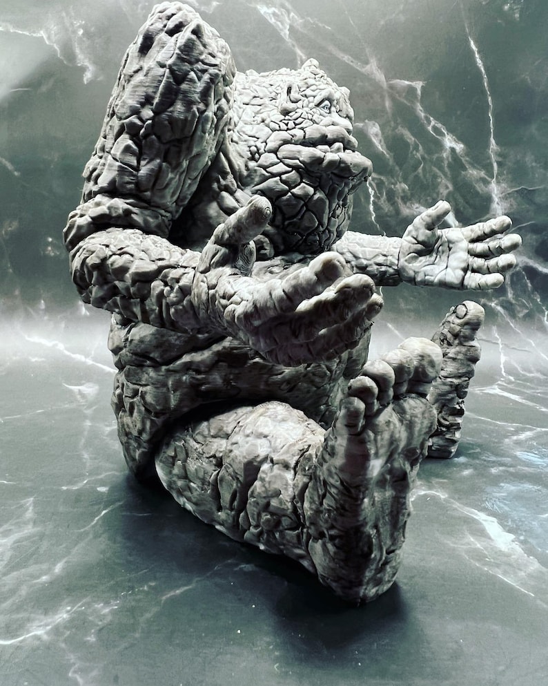 May include: A grey stone sculpture of a creature with a textured surface. The creature is sitting with its legs crossed and its arms outstretched.
