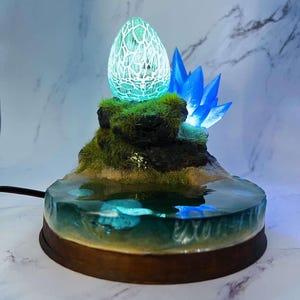 Può includere: Un uovo di drago blu incandescente si trova su una base di roccia ricoperta di muschio con un cristallo blu accanto. La base è circondata da una piscina di resina trasparente con un fondo sabbioso.