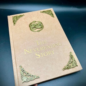 Puede incluir: Un libro de tapa dura color canela con el título The Neverending Story en letras doradas. La portada presenta un símbolo Auryn dorado de dos serpientes entrelazadas y adornos metálicos en las tres esquinas visibles del libro.
