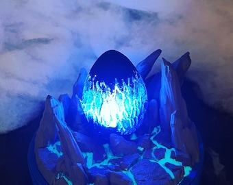 May include: A blue and white 3D printed dragon egg with a glowing blue light inside, sitting on a rocky base with blue accents.