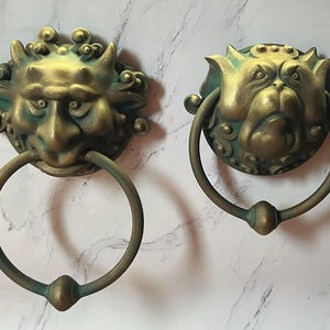 May include: Two antique-style door knockers. Each knocker features a detailed, sculpted face with a ring handle. The faces have a bronze finish with green accents, and the rings are attached to the face with a decorative base. The knockers are mounted on a white marble-like surface.