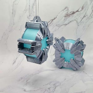 May include: Two futuristic-looking ornaments. Each ornament has a light blue center and is encased in a silver, geometric design. The ornaments are suspended by thin silver strings, suggesting they are designed for hanging. The overall aesthetic is modern and sci-fi.