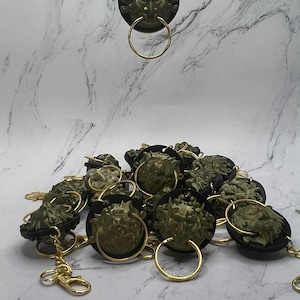 May include: Keyrings with a detailed green face design, gold-coloured ring, and chain. The face is set against a black circular background. The keyrings are scattered on a marble-effect surface. The keyrings are a unique accessory.
