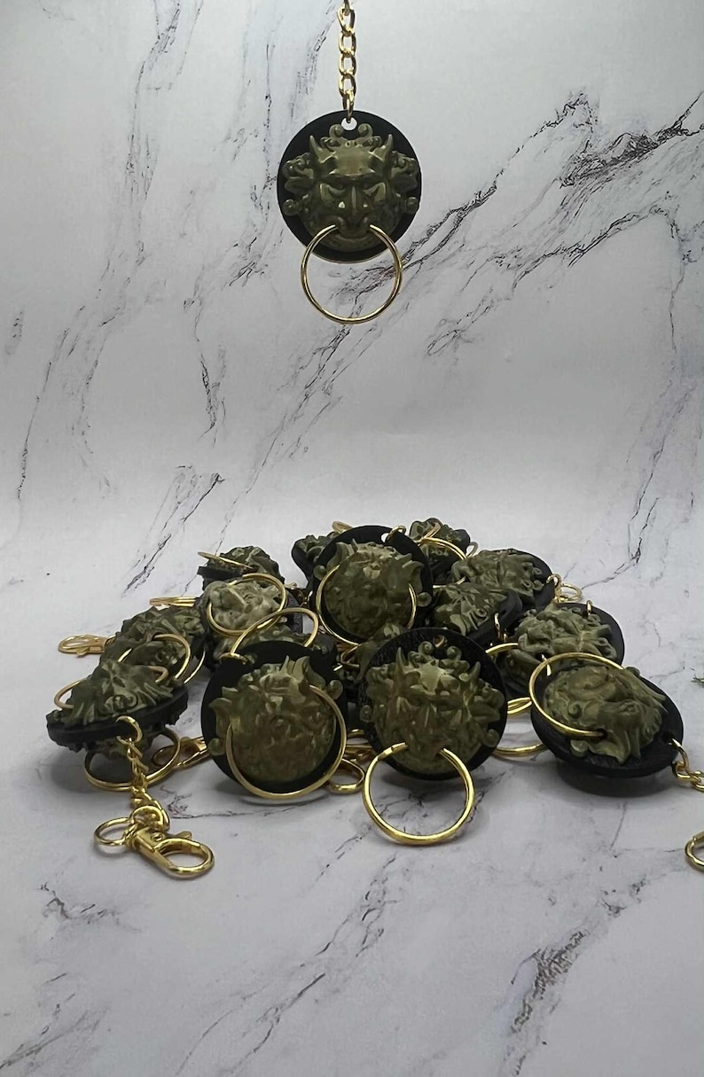 May include: Keyrings with a detailed green face design, gold-coloured ring, and chain. The face is set against a black circular background. The keyrings are scattered on a marble-effect surface. The keyrings are a unique accessory.
