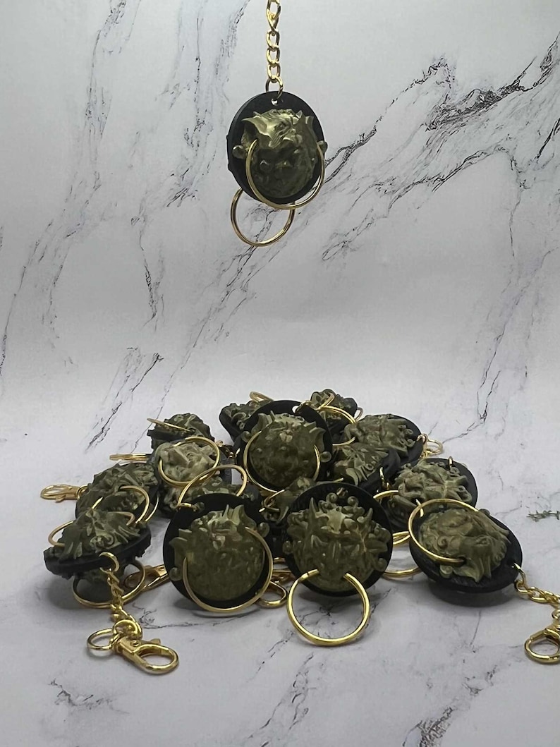 May include: Keyrings with a lion's head design. Each keyring has a gold-coloured chain and ring, with a dark circular backing. The lion's head is a muted green colour.