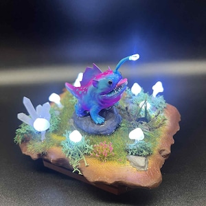 May include: A fantasy sculpture of a blue and purple anglerfish with a glowing lure. The fish is set on a miniature landscape with glowing mushrooms, crystals, and moss. The base is a wooden-like structure.