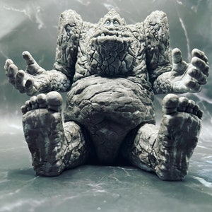 May include: A grey stone creature with a wide, toothy grin sits with its legs crossed and arms outstretched. The creature has a textured, rocky surface and appears to be made of stone.