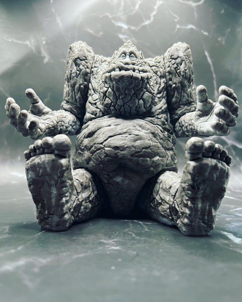 May include: A grey stone creature with a wide, toothy grin sits with its legs crossed and arms outstretched. The creature has a textured, rocky surface and appears to be made of stone.
