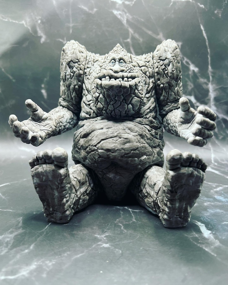 May include: A grey stone figure with a wide smile and large feet. The figure is sitting with its legs crossed and its arms outstretched.
