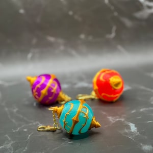 May include: Three decorative ornaments with gold accents. The ornaments are round and have a pointed tip. The ornaments are blue, purple, and red.
