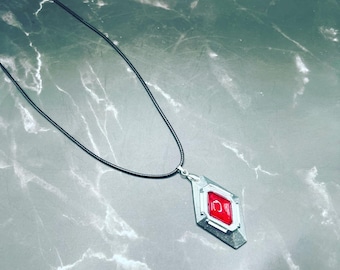 May include: A silver pendant necklace with a red gemstone in the center. The pendant is shaped like a diamond and has a black cord.