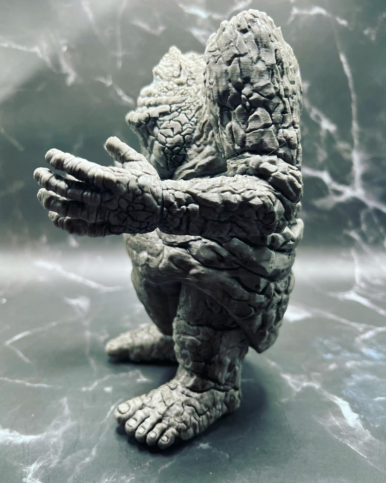 May include: A grey stone-like figure with a textured surface. The figure is in a crouching position with its arms raised.