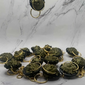 May include: Bronze-coloured keychains with a sculpted face design. Each keychain has a black backing, a gold-coloured ring, and a gold-coloured chain. The keychains are arranged on a white and grey marble-patterned surface.