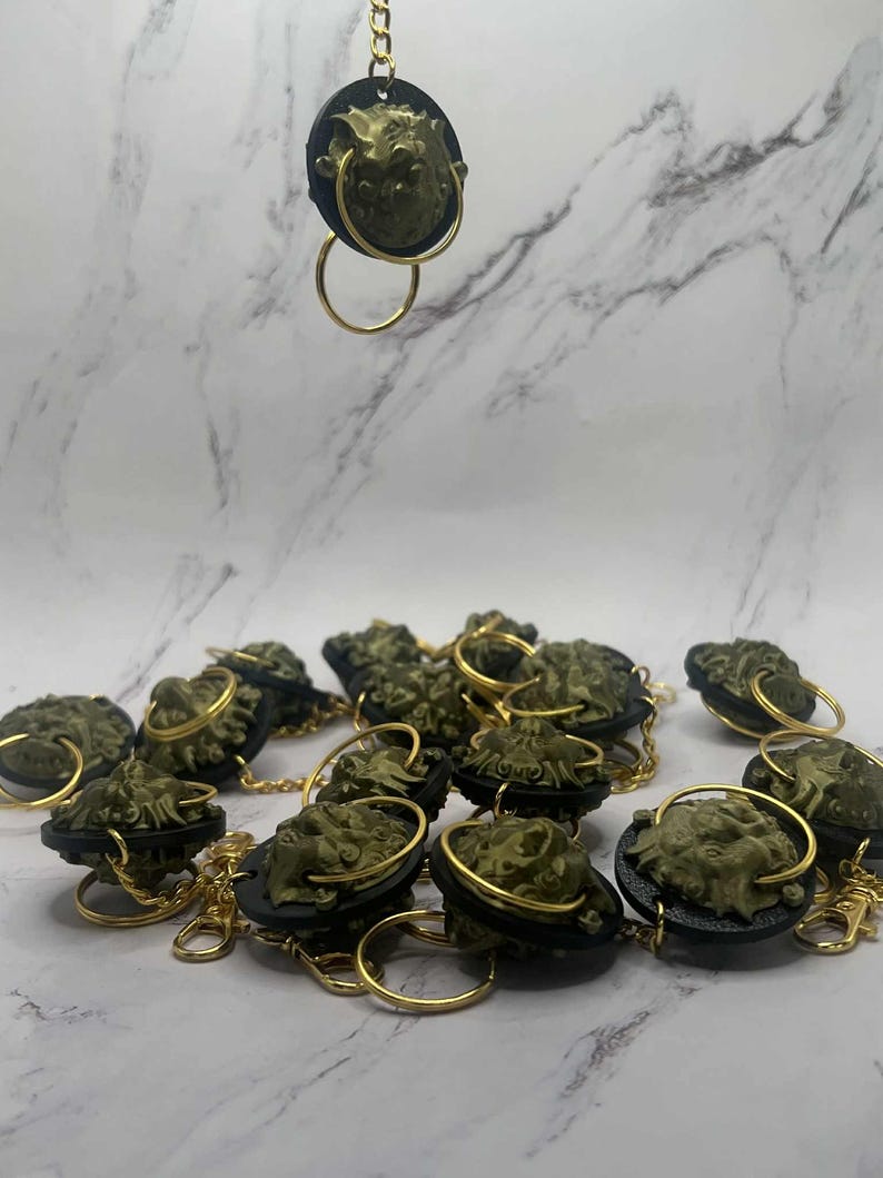 May include: Bronze-coloured keychains with a sculpted face design. Each keychain has a black backing, a gold-coloured ring, and a gold-coloured chain. The keychains are arranged on a white and grey marble-patterned surface.