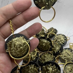 May include: A selection of gold-coloured lion head keychains. Each keychain features a detailed lion head design attached to a black circular base, with a gold ring and chain. The keychains are scattered on a white marble surface.