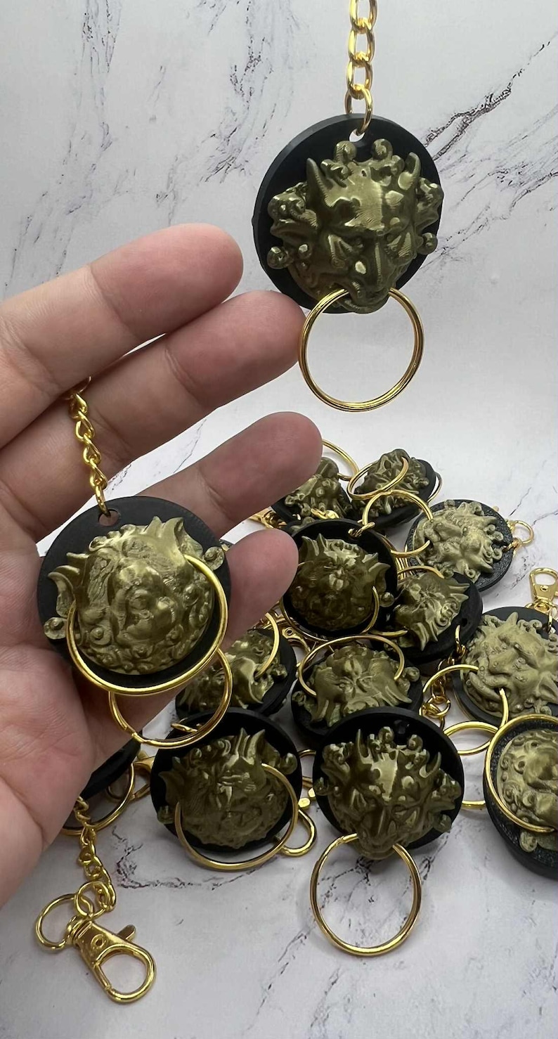 May include: A selection of gold-coloured lion head keychains. Each keychain features a detailed lion head design attached to a black circular base, with a gold ring and chain. The keychains are scattered on a white marble surface.
