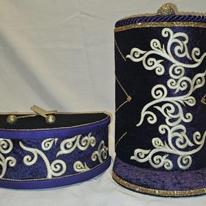 May include: A deep purple velvet paper towel holder and a matching semi-circular storage container. Both items feature intricate white embroidered scrollwork, gold-toned trim, and a small gold-toned ball finial on the top of the towel holder.