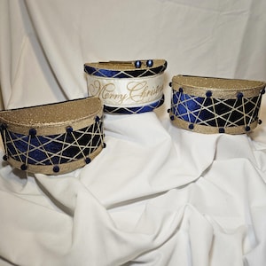 Op de afbeelding: Drie halfronde, trommelvormige ornamenten met donkerblauwe fluwelen zijkanten, gouden kruislings geplaatst koord en kleine blauwe pompons. De bovenranden zijn bedekt met gouden glitters. Eén ornament bevat de tekst Merry Christmas in goud.