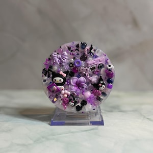 Könnte beinhalten: Ein rundes, dekoratives Harzstück mit klarer Basis, verziert mit verschiedenen lila und schwarzen Verzierungen. Das Design umfasst Schleifen, Blumen, Perlen und Cartoon-Charakterfiguren. Das Stück ist auf einem transparenten Ständer montiert.