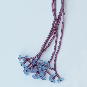 Puede incluir: Un delicado collar con un grupo de dijes de flores azul claro. El collar tiene una hebra central de cuentas moradas que se divide en múltiples hebras. El collar está adornado con pequeños dijes de flores azul claro.