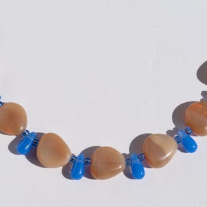 May include: A necklace featuring large, peach-colored, heart-shaped beads interspersed with small, bright blue beads. The necklace is displayed against a white background, highlighting the contrast of colors and shapes.