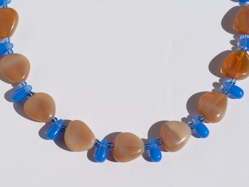 May include: A necklace featuring large, peach-colored, heart-shaped beads interspersed with small, bright blue beads. The necklace is displayed against a white background, highlighting the contrast of colors and shapes.