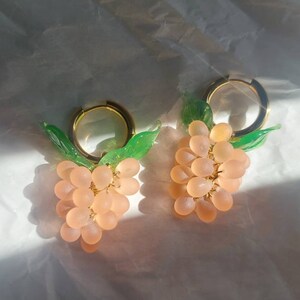 Puede incluir: Un par de pendientes de aro dorados con uvas de cristal rosa y hojas verdes que cuelgan.