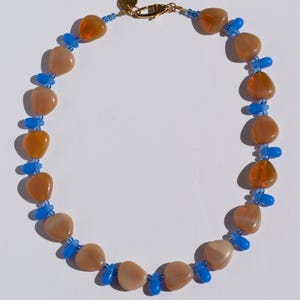 May include: A necklace featuring a mix of peach and orange-colored, irregularly shaped beads, interspersed with small blue beads. The necklace has a gold-colored clasp and a small gold charm.