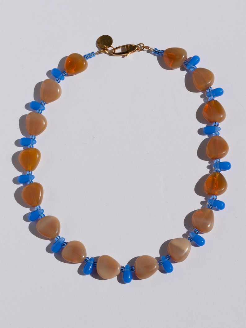 May include: A necklace featuring a mix of peach and orange-colored, irregularly shaped beads, interspersed with small blue beads. The necklace has a gold-colored clasp and a small gold charm.