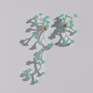 Puede incluir: Un par de pendientes plateados con un diseño floral. Cada pendiente presenta una estructura ramificada con pequeñas cuentas en forma de flor de color azul claro. Los pendientes se exhiben sobre una superficie blanca, proyectando sombras.