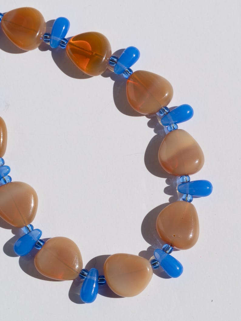 May include: A necklace featuring large, rounded, peach-colored beads interspersed with bright blue, teardrop-shaped beads. The necklace is strung on a clear cord, creating a striking contrast between the colors and shapes. A unique piece of jewelry.