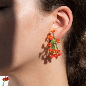 May include: Floral earrings featuring clusters of small red flowers with yellow centers and green stems. The earrings are worn on the ear, with the shadow of the earrings visible on the skin. The earrings are a unique and eye-catching accessory.