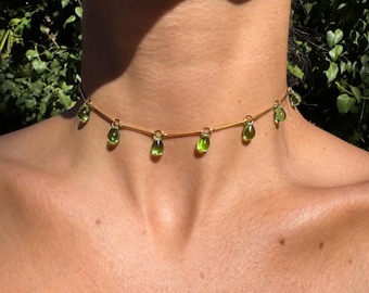 Puede incluir: Un delicado collar gargantilla dorado con piedras preciosas verdes en forma de lágrima. El collar está ensartado en una fina cadena dorada, con las piedras espaciadas en la parte delantera. Una joya sencilla pero elegante.