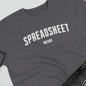 Puede incluir: Una sudadera gris oscuro con la inscripción blanca "SPREADSHEET WIZARD" en arco. La imagen también muestra unos vaqueros oscuros, un cinturón blanco y zapatillas deportivas blancas y negras.