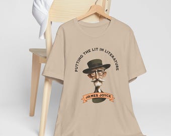 May include: Beige t-shirt featuring a cartoon illustration of James Joyce with a hat and mustache. The text "PUTTING THE LIT IN LITERATURE" and "JAMES JOYCE" are also printed on the shirt. A white baseball cap rests on a wooden chair.