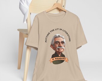 May include: Beige t-shirt featuring a cartoon illustration of a man with glasses and a mustache. The text "PUTTING THE LIT IN LITERATURE" is arched around the illustration. Below the illustration is a banner with the text "G. MARQUEZ". A white baseball cap rests on a wooden chair.