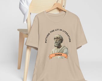 May include: Beige t-shirt with a graphic of Homer, a Greek poet, and the text "Putting the Lit in Literature." The shirt is hanging on a wooden hanger. A white baseball cap rests on a wooden chair.