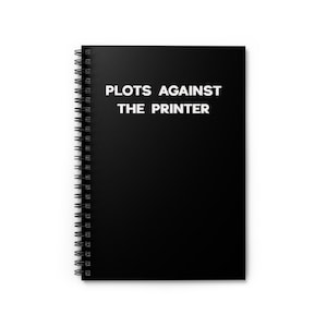 May include: A black spiral-bound notebook with the text "PLOTS AGAINST THE PRINTER" in white, centered on the cover. The notebook's pages are white, and the spiral binding is silver.